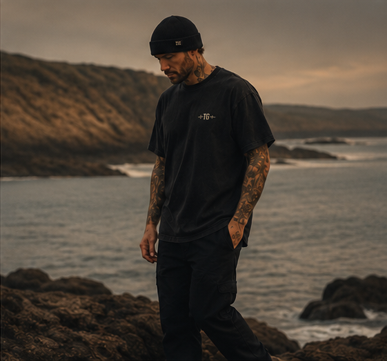 Full-length editorial shot of a man in a black Tinnitus Gang tee and beanie standing on coastal rocks